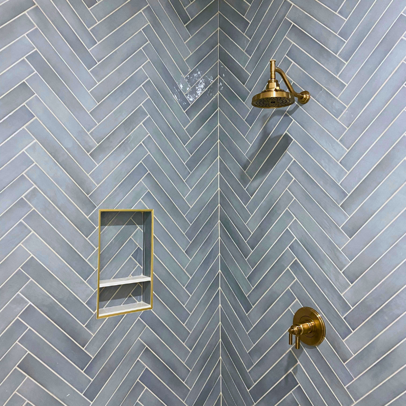 Light blue herringbone tile installed in a luxurious shower with gold fixtures and a well-crafted niche.